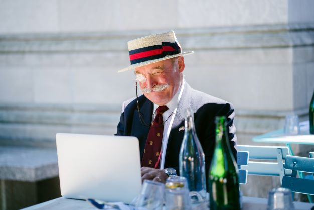 Elder gentleman in suit and summerhat sitting outside at a coffeetable with a laptop, smiling looking amused.